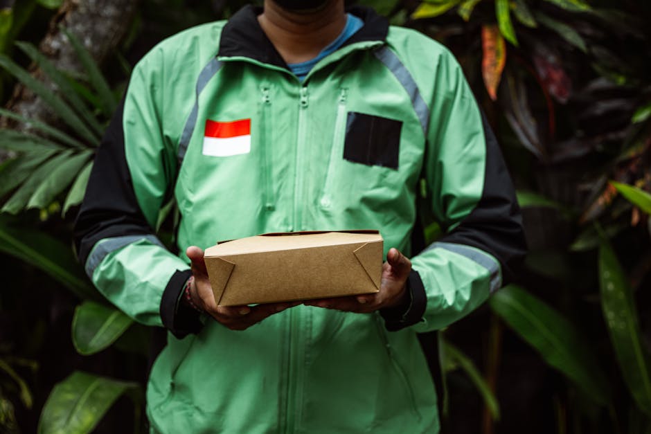 A delivery person wearing a bright green jacket with black and reflective grey accents, holds a rectangular cardboard box with folded edges in both hands, positioned at waist level. The jacket feature